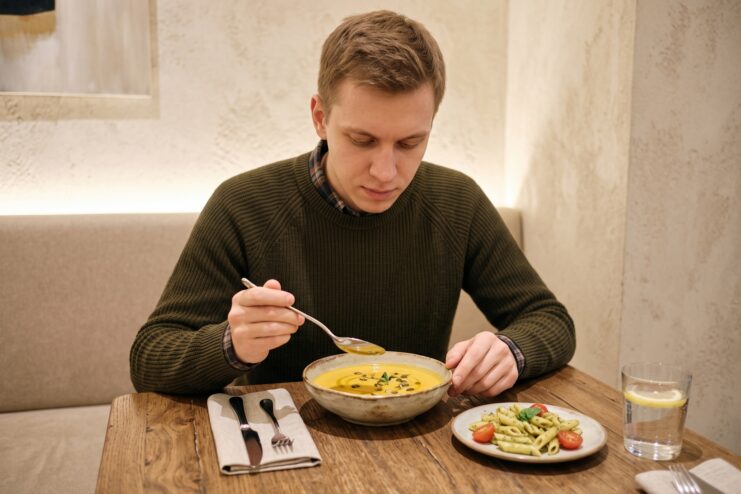 Consumo de comida sencilla y cómoda durante la ansiedad Hombre comiendo sopa con cuchara en la mesa de un café, junto a un platillo de pasta penne.