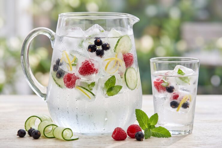 Jarra de cristal y vaso de agua con hielo que contiene bayas, pepino y menta en su interior.